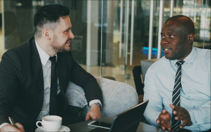 Two colleagues talking over coffee at a table in a bright office.